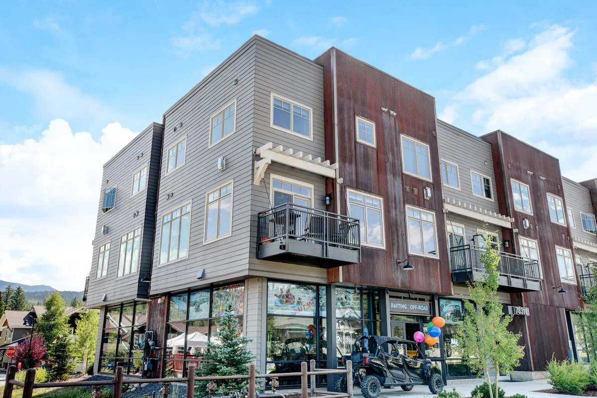 A lively exterior scene showcasing modern architecture, balconies adorned with colorful balloons, and people enjoying the vibrant atmosphere. Greenery frames the walkways, while groups are seen shopping, socializing, and dining outdoors, suggesting a festive event or regular community gathering.