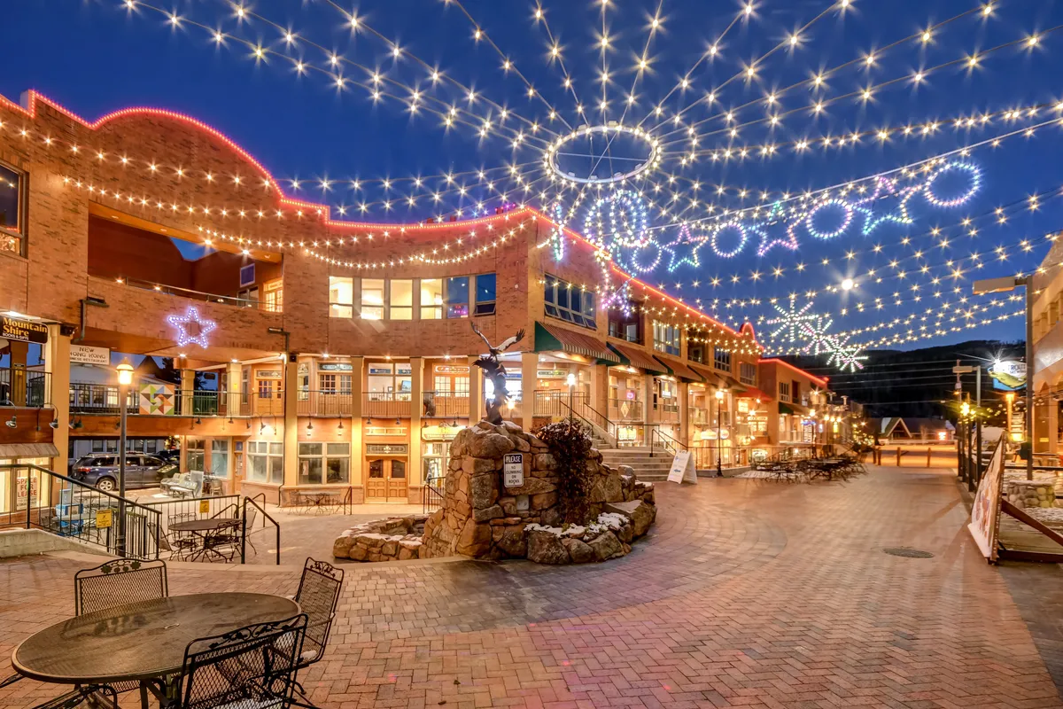 An inviting exterior shot of a festive plaza, adorned with string lights and decorative stars. The stone fountain and nearby shops highlight a lively community setting, ideal for socializing, dining, and family outings.