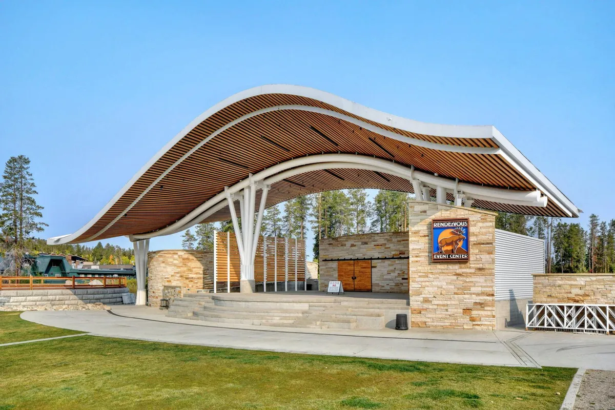 An exterior view of a unique curved roof structure with a stone facade and wooden architectural details, serving as an amphitheater. The setting evokes the site of concerts and community celebrations, with ample space for audiences to gather outdoors.