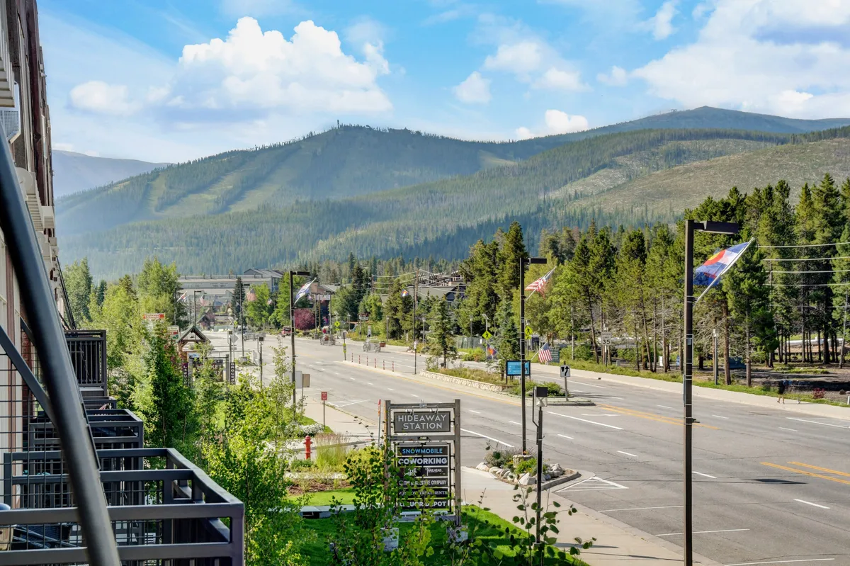 A serene and inviting view of Winter Park’s mountain range, with the bustling Main Street, trees, flags, and the Hideaway Station sign in the foreground. The scene captures the essence of the town, highlighting opportunities for hiking, biking, and immersion in the local community.