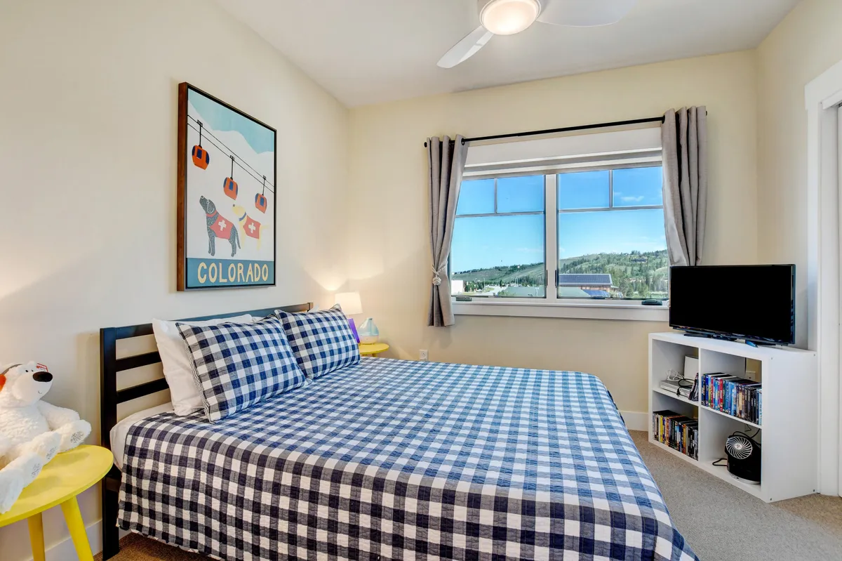 A cozy bedroom with plaid bedding and Colorado-themed artwork, bathed in natural light from a large window overlooking the mountains. The atmosphere is warm and inviting, ideal for relaxing after a day of urban adventure or mountain exploration.