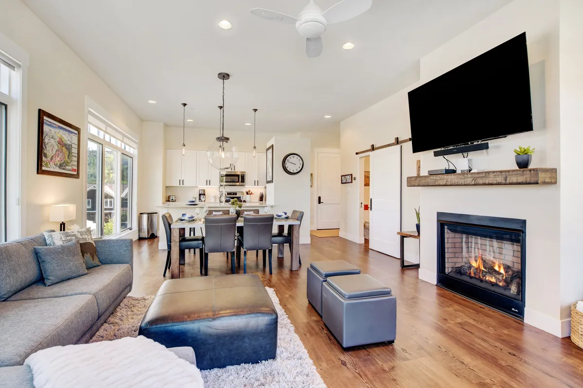 The image showcases a warm and inviting open-concept living space featuring a plush sofa, a dining table perfect for gatherings, a modern fireplace, and a large flat-screen television. Soft lighting and tasteful mountain-inspired decor create a cozy atmosphere, ideal for relaxing or entertaining after a day of Colorado adventures.