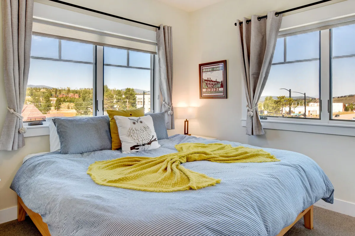 A cozy bedroom with a plush bed layered in premium bedding, a large window offering a scenic view, a decorative pillow, and a warm yellow blanket. The soft lighting and inviting atmosphere suggest a restful retreat.
