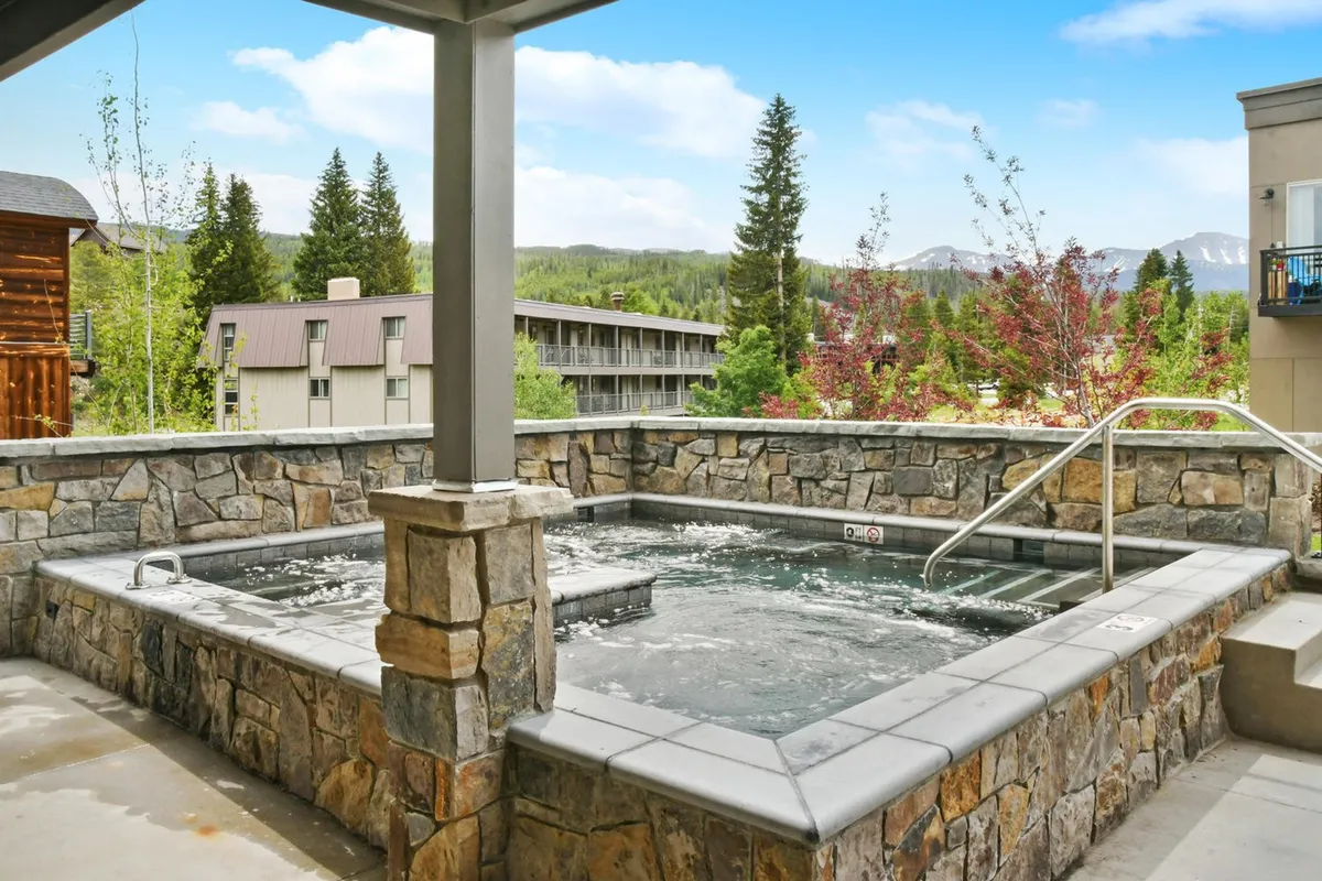 An expansive outdoor hot tub surrounded by lush greenery and set against a stunning mountain backdrop. Several guests are relaxing in the steaming water, enjoying the tranquil natural setting, while others socialize nearby.