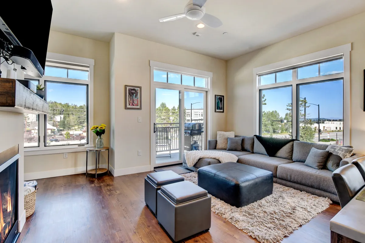 A bright, inviting living area with a plush sofa and coffee table set in front of a modern fireplace. Floor-to-ceiling windows and a glass door open onto a balcony, allowing natural light to pour in and offering views of the outdoors. The stylish décor and welcoming mood make it a perfect spot for family games or quiet reading.