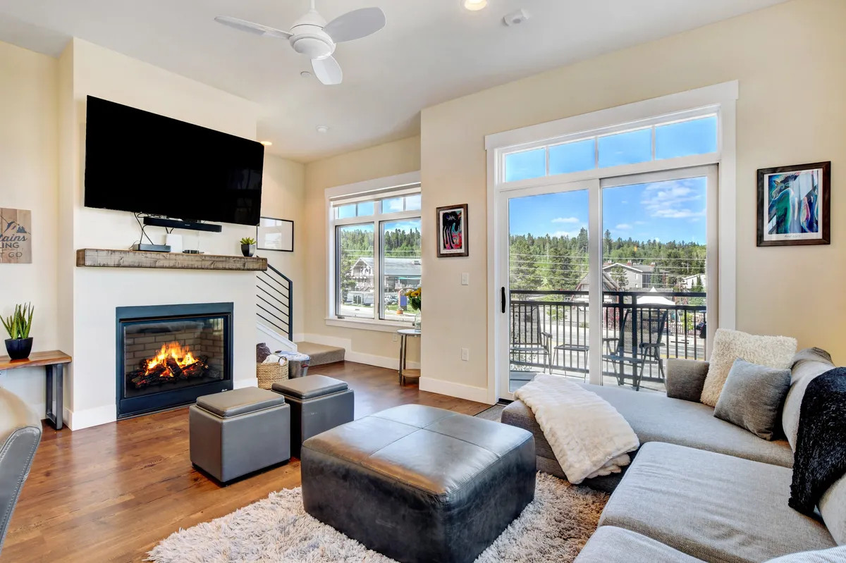 A spacious living room featuring a comfortable sofa arranged around a modern, programmable gas fireplace. Large windows and glass doors open to a balcony with scenic views of the surrounding mountains and downtown, creating a seamless connection between the indoor living space and outdoor beauty. The room exudes coziness and is ideal for relaxing or entertaining guests.
