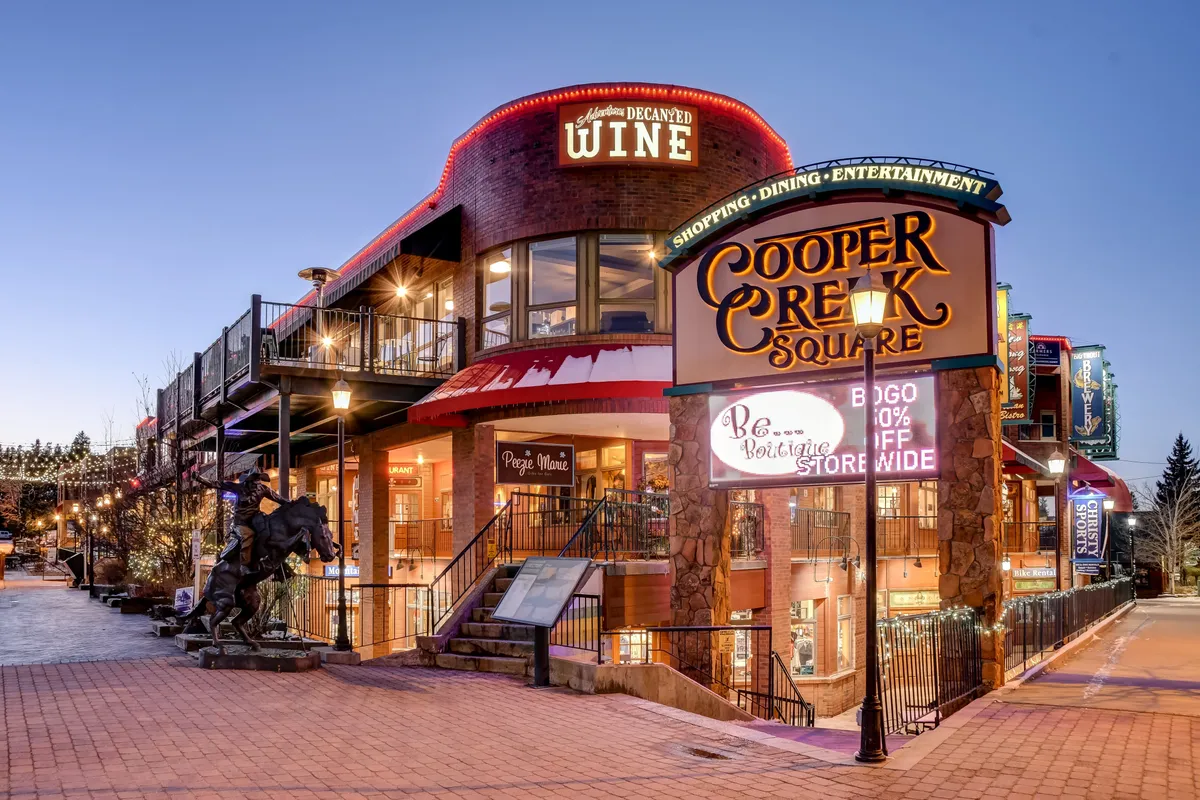 A bustling streetscape featuring the Cooper Creek Square sign, a wine shop, a staircase, and a striking horse sculpture. The area feels lively and festive, indicative of a hub for shopping, dining, and entertainment, with hints of seasonal or special events through decorations.