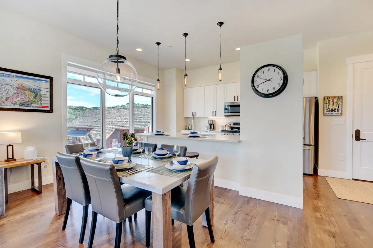 A bright, well-appointed dining area adjoining the kitchen, featuring a spacious dining table set near a large window. The clock and homey details create a scene ideal for shared meals, celebrations, or quiet mornings with coffee.